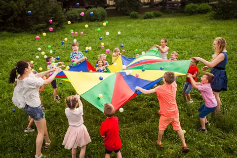 Un groupe d'enfants en train de jouer dans un pré avec une grande couverture et des balles en plastiques qu'ils font sauter dessus